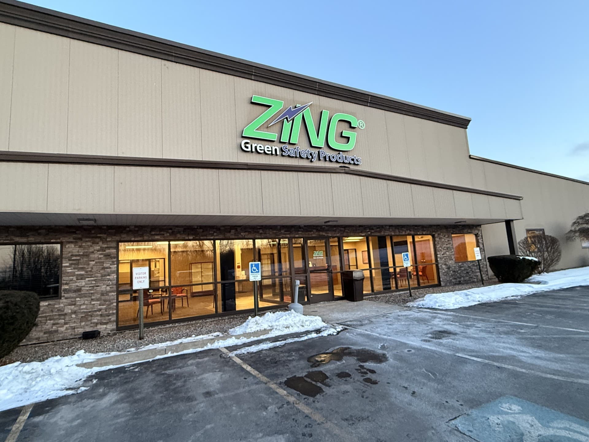 Exterior of a commercial building with a "ZING Green Safety Products" sign. Snow on the ground, accessible parking spaces visible.